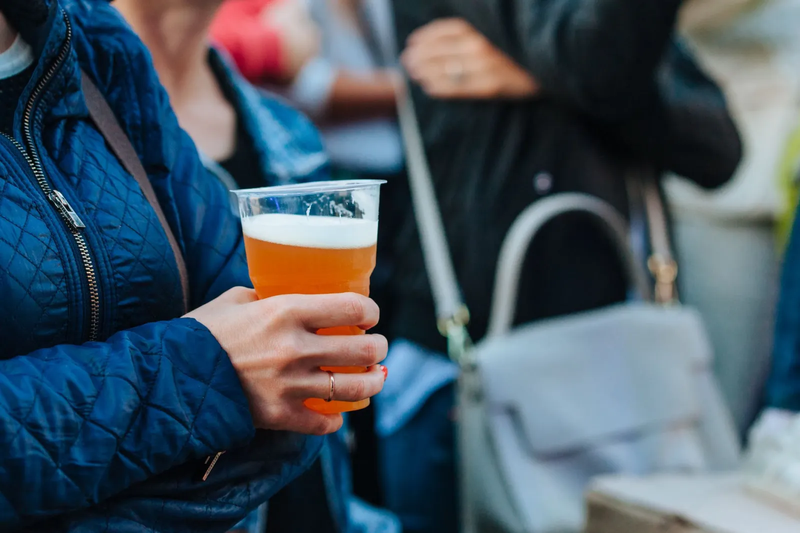 Osoba trzymająca kubek z próbką piwa na zewnętrznym festiwalu piwnym, w tle tłum i stoiska